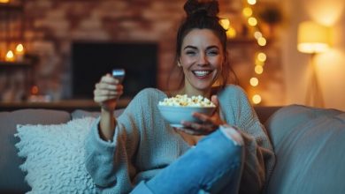 jovem relaxada deitada em um sofa com um controle remoto em uma mao e uma tigela de pipoca na outra sorrindo para a camera 132358 65888