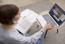 Mulher participando de um curso online, assistindo a uma aula em vídeo enquanto estuda com um livro aberto