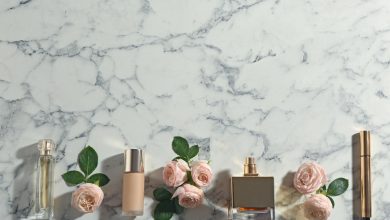 Frascos de perfume, base de maquiagem e rosas cor de rosa dispostos em fileira sobre superfície de mármore branco