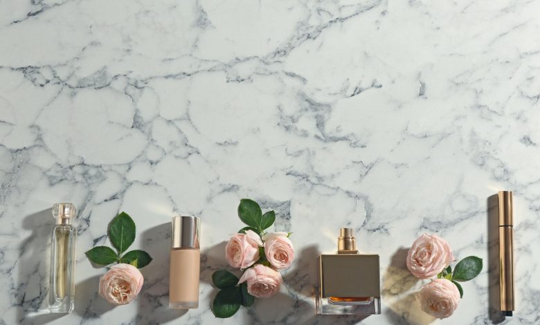 Frascos de perfume, base de maquiagem e rosas cor de rosa dispostos em fileira sobre superfície de mármore branco